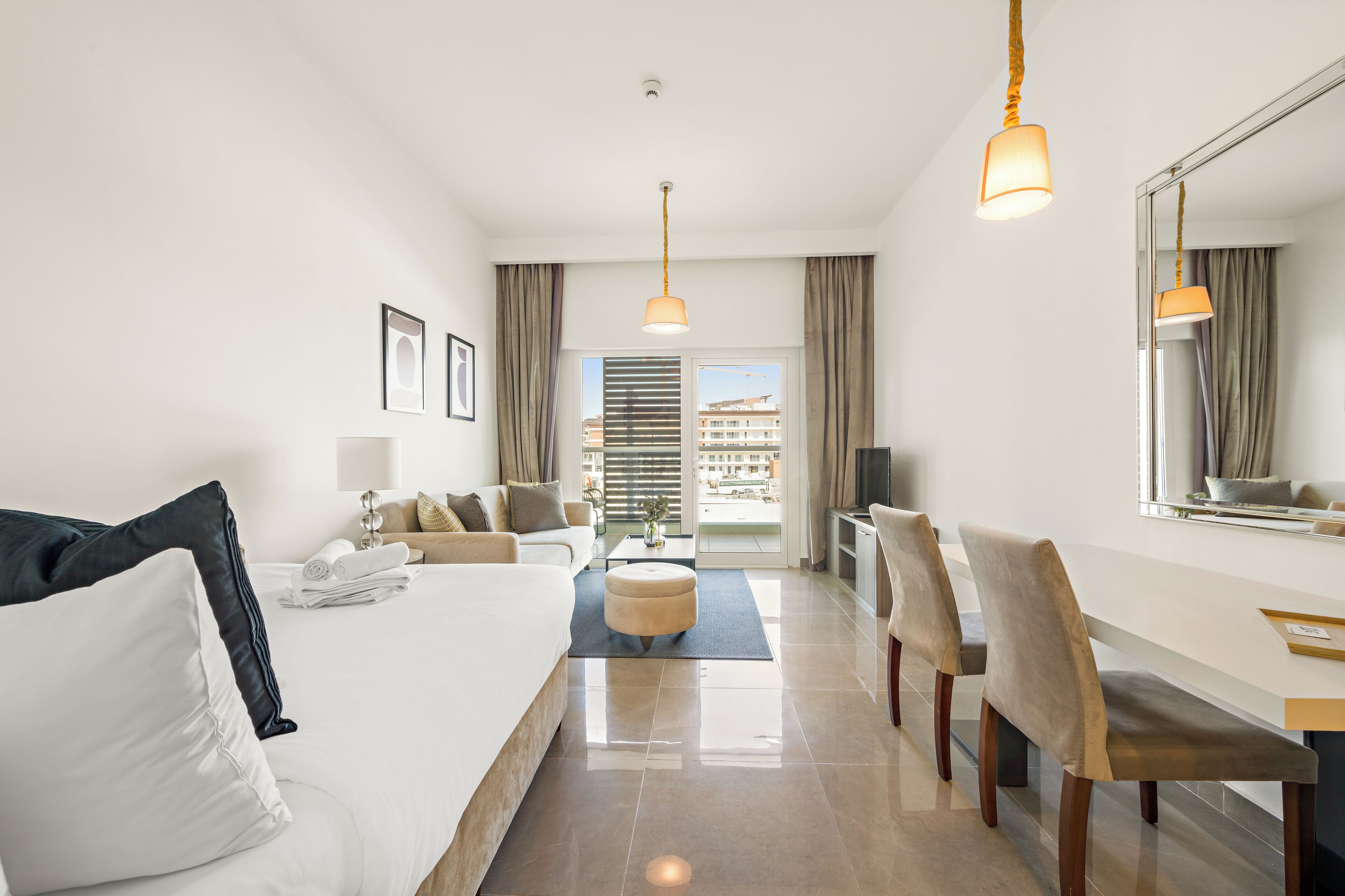 A hotel room with a bed, a sofa, a dining table, chairs, a TV, and a large window with a view of buildings.