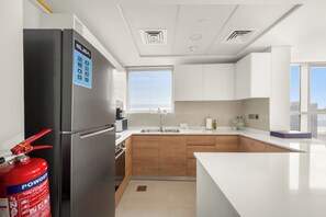 A modern kitchen with a stainless steel refrigerator, wooden cabinets, a white countertop, and a window with blinds. - Silkhaus Radiant Heights (Abu Dhabi)