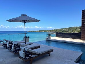 A poolside area with lounge chairs and an umbrella, overlooking a clear blue sea and a hilly landscape.