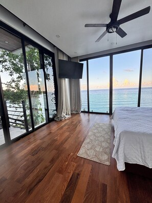 A modern bedroom with a large bed, a ceiling fan, and a view of the sea.