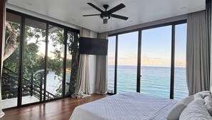 Ett sovrum med en stor säng, en takfläkt och en tv. Det har stora fönster med utsikt över havet.