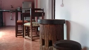 Ein Zimmer mit Holzboden, einem kleinen Tisch, einem Stuhl und einem Hocker. Es gibt ein Fenster mit Vorhang und eine Wandleuchte.