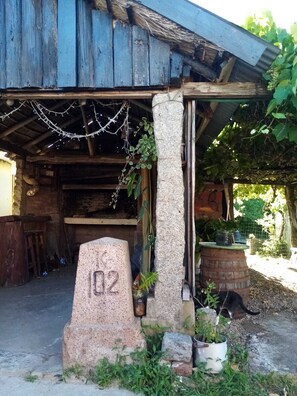 Un bâtiment rustique avec un pilier en pierre portant le numéro « 102 », des poutres en bois et un espace extérieur abrité agrémenté de plantes en pot.