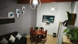 Een gezellig, compact ingerichte ruimte met een zwarte bank, een houten eettafel en stoelen. Een flatscreen televisie is aan de muur bevestigd, en er hangen verschillende kunstwerken boven de tafel.