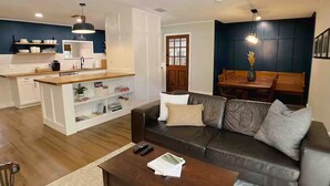 Un salon moderne avec un canapé en cuir foncé, une table basse en bois et un coin cuisine doté de meubles blancs et d’un plan de travail en bois.