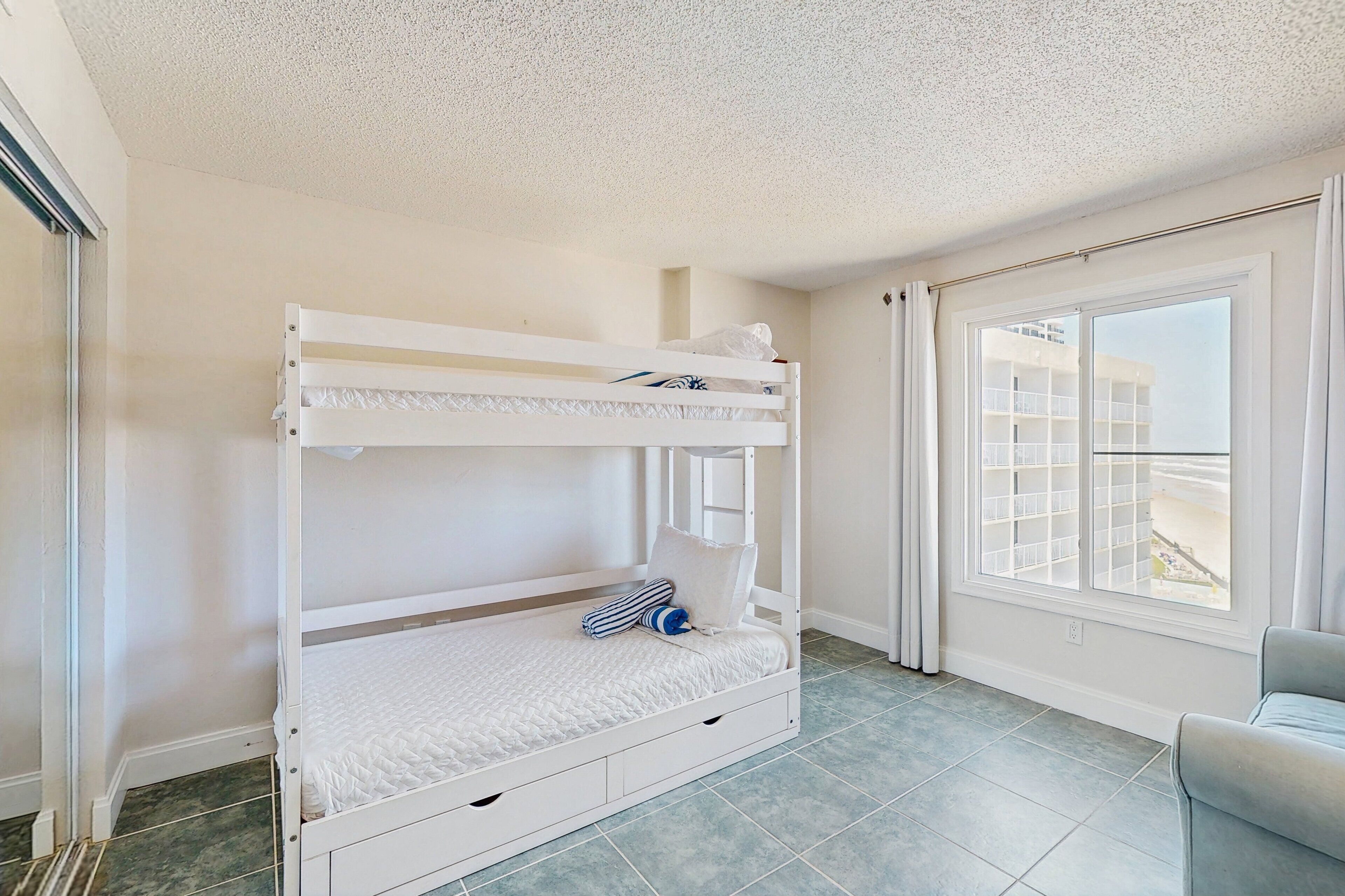 A bunk bed with a white mattress and pillows, a grey chair, and a window with curtains.