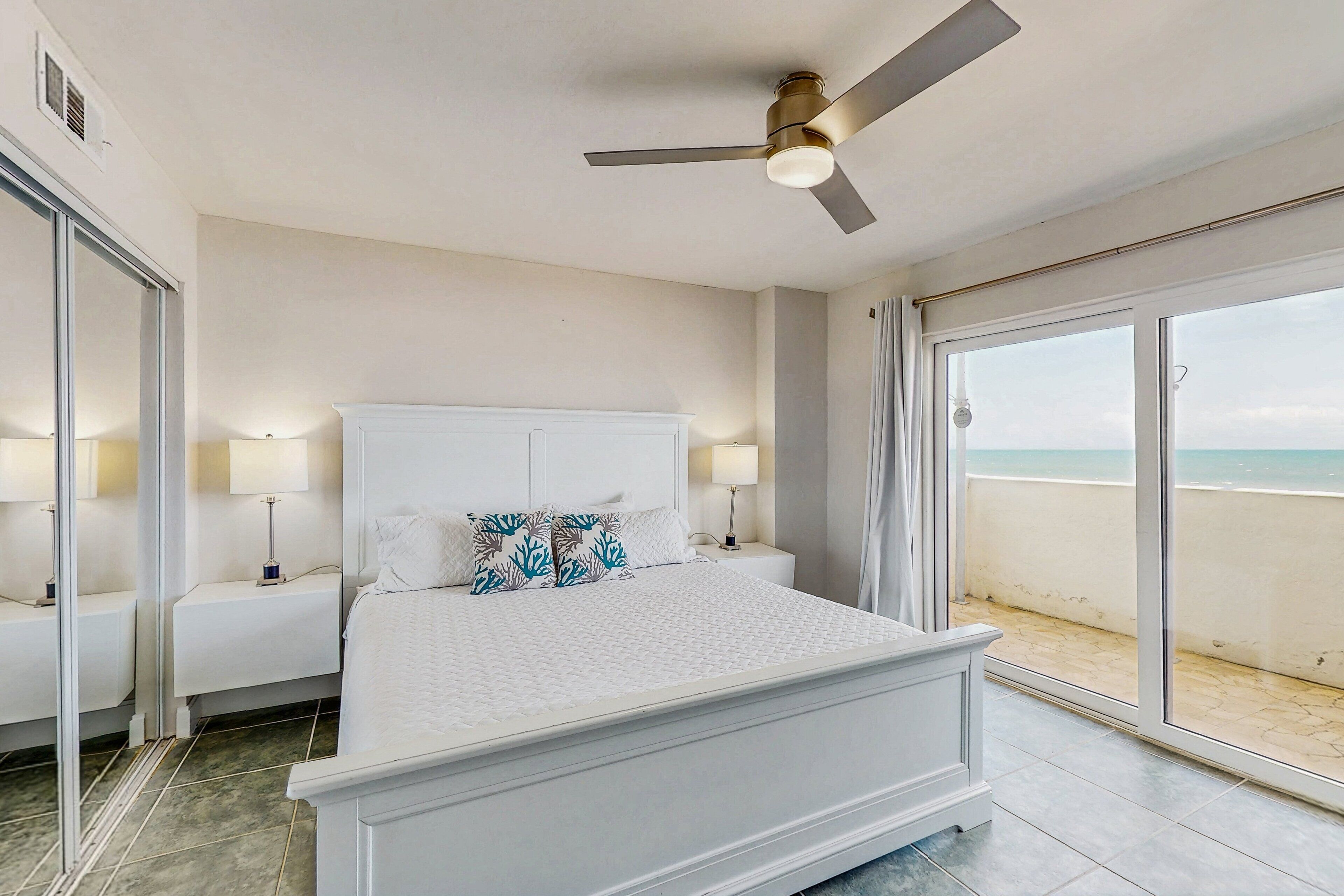 A bedroom with a bed, nightstands, a ceiling fan, and a view of the beach through large sliding glass doors.