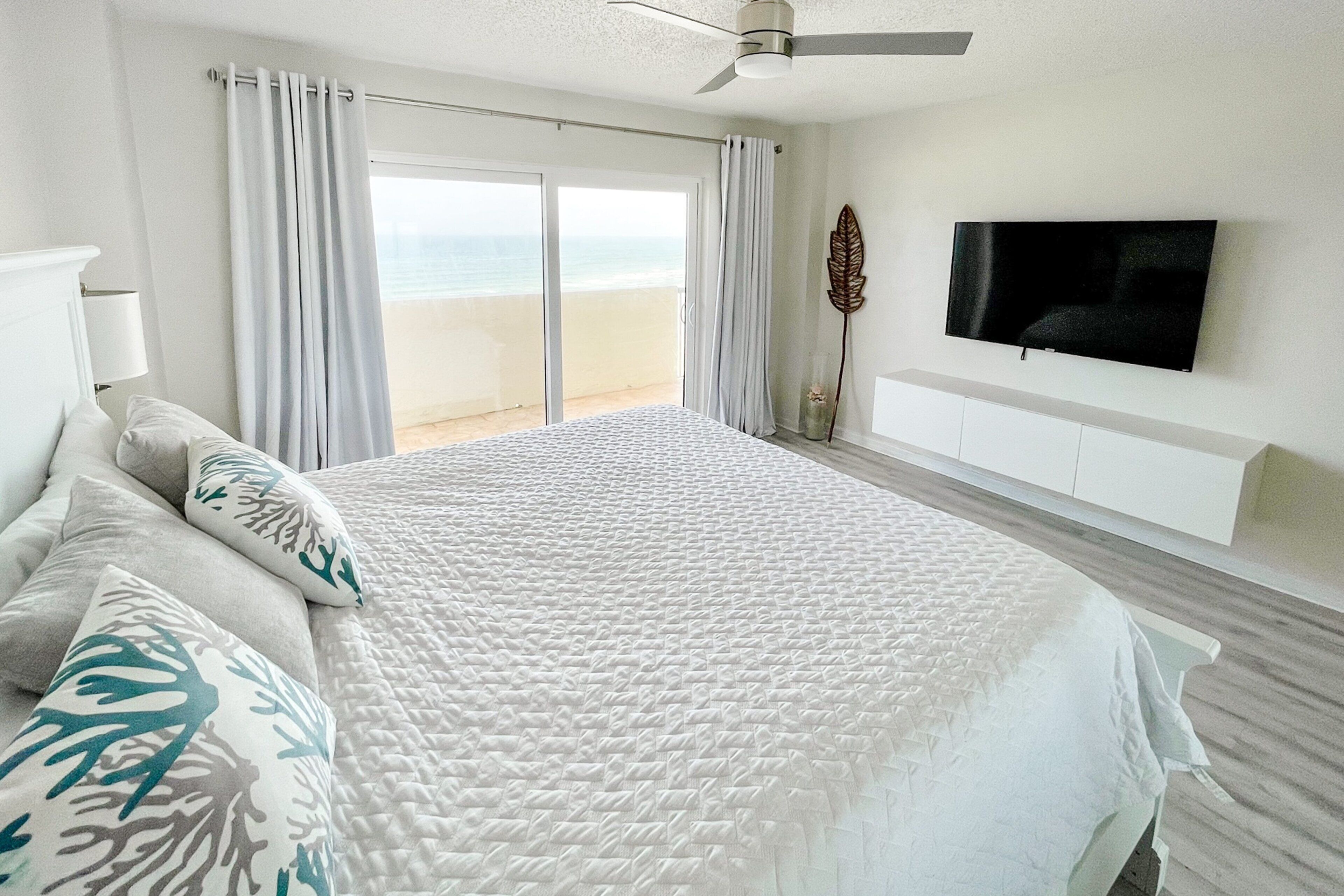 A bedroom with a large bed, a ceiling fan, a TV mounted on the wall, and a view of the beach through the sliding glass doors.