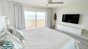 Ein Schlafzimmer mit einem großen Bett, einem Deckenventilator, einem an der Wand befestigten Fernseher und Blick auf den Strand durch Glastüren.