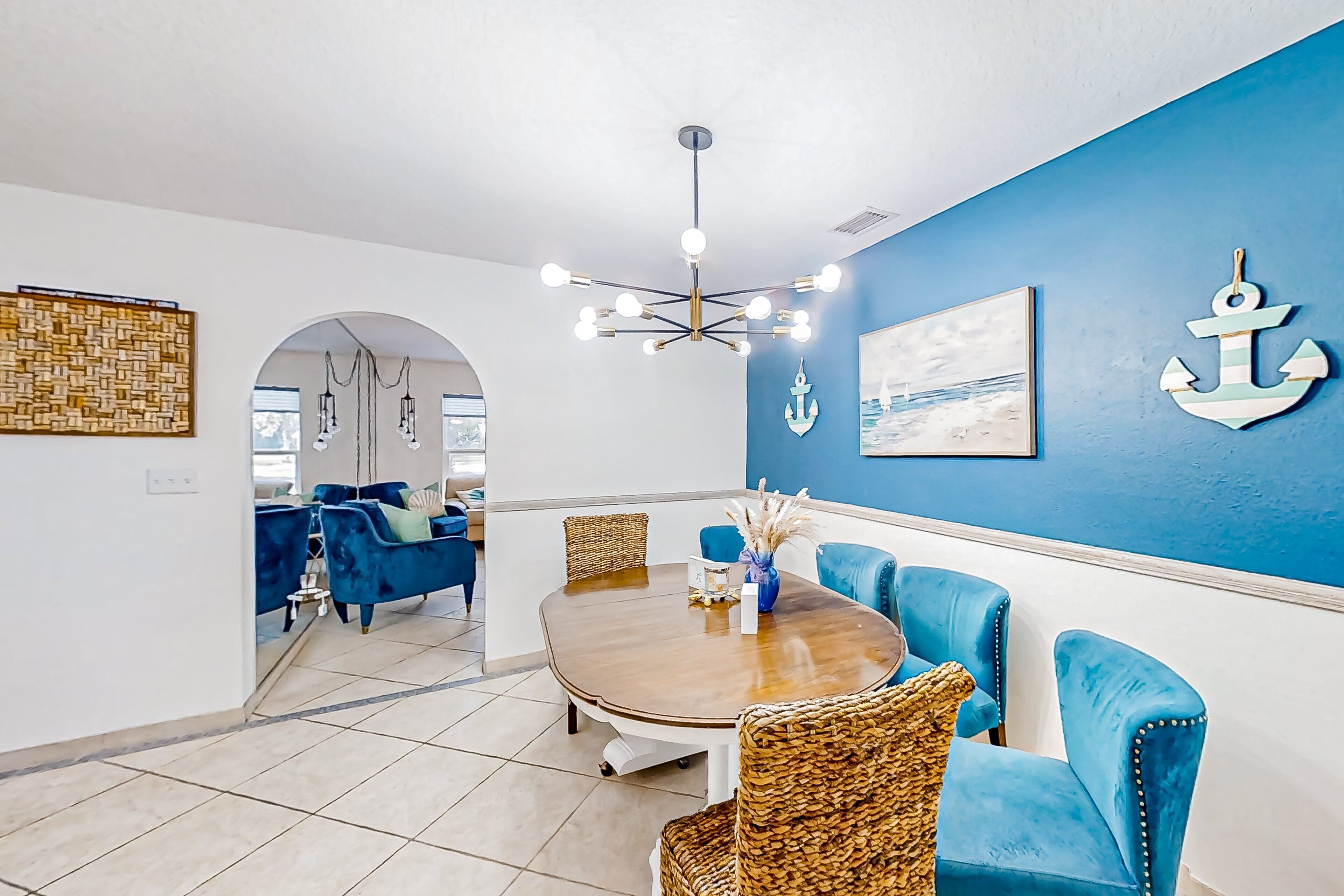 A dining area with a round table, blue upholstered chairs, a hanging light fixture, and nautical-themed wall decor.