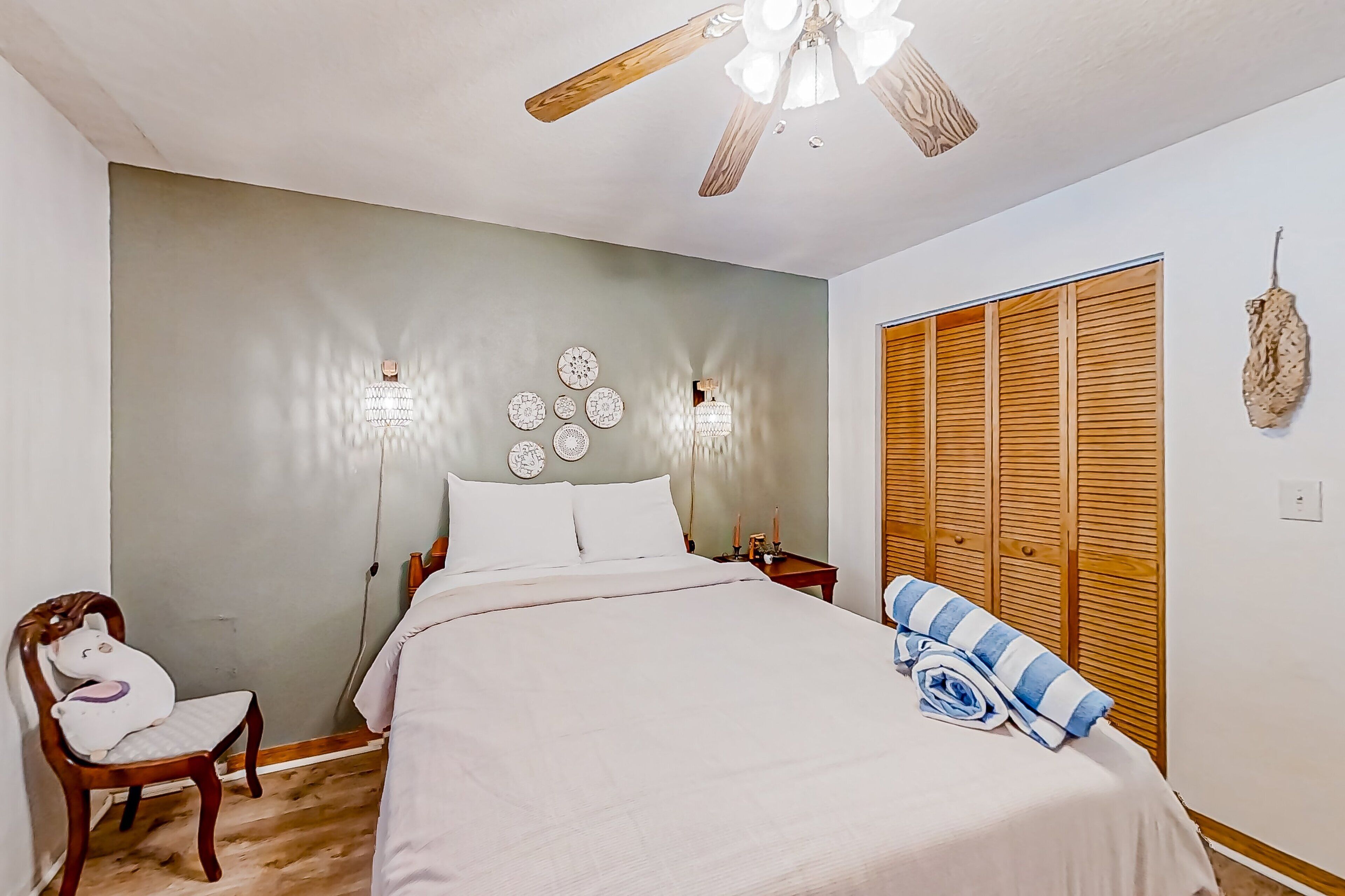Un dormitorio con cama, una silla de madera, ventilador de techo, apliques de pared y un armario de madera.