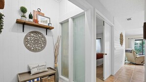 A hallway with a wooden bench, wicker baskets, a welcome home pillow, decorative wall art, and a glass door leading to another room. - NSB 4360 Sea Woods- After Dune Delight (New Smyrna Beach)