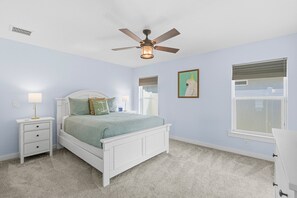 A bedroom with a white bed, a nightstand, a ceiling fan, a window with blinds, and a framed picture on the wall. - Terrapin Station (Ormond Beach)