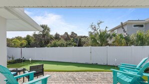 A patio with turquoise Adirondack chairs, a brick floor, and a view of a backyard with a white fence and greenery. - Terrapin Station (Ormond Beach)