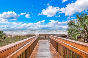 Un paseo marítimo de madera que conduce a una playa con cielo despejado y nubes.