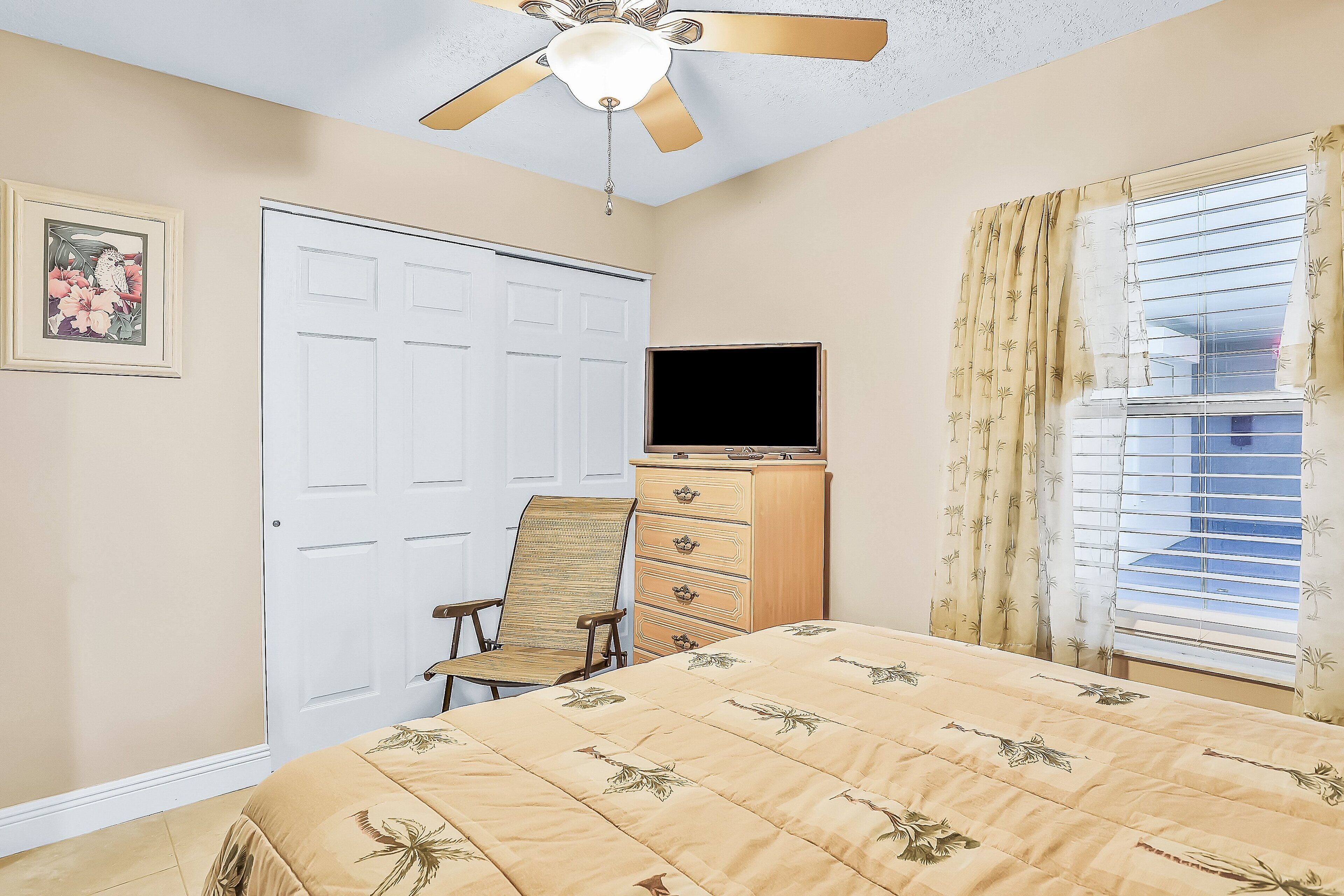 A bedroom with a bed, a chair, a dresser, a television, a ceiling fan, and a window with curtains.