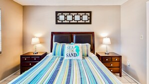 A neatly made bed with striped bedding and a pillow with a coastal design, flanked by two bedside tables with lamps. - NSB Seaward D-101 (New Smyrna Beach)