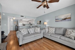 A living room with two grey sofas, patterned cushions, a ceiling fan, and a kitchen in the background. - Ormondy A6 - Ocean Shores Getaway (Ormond Beach)