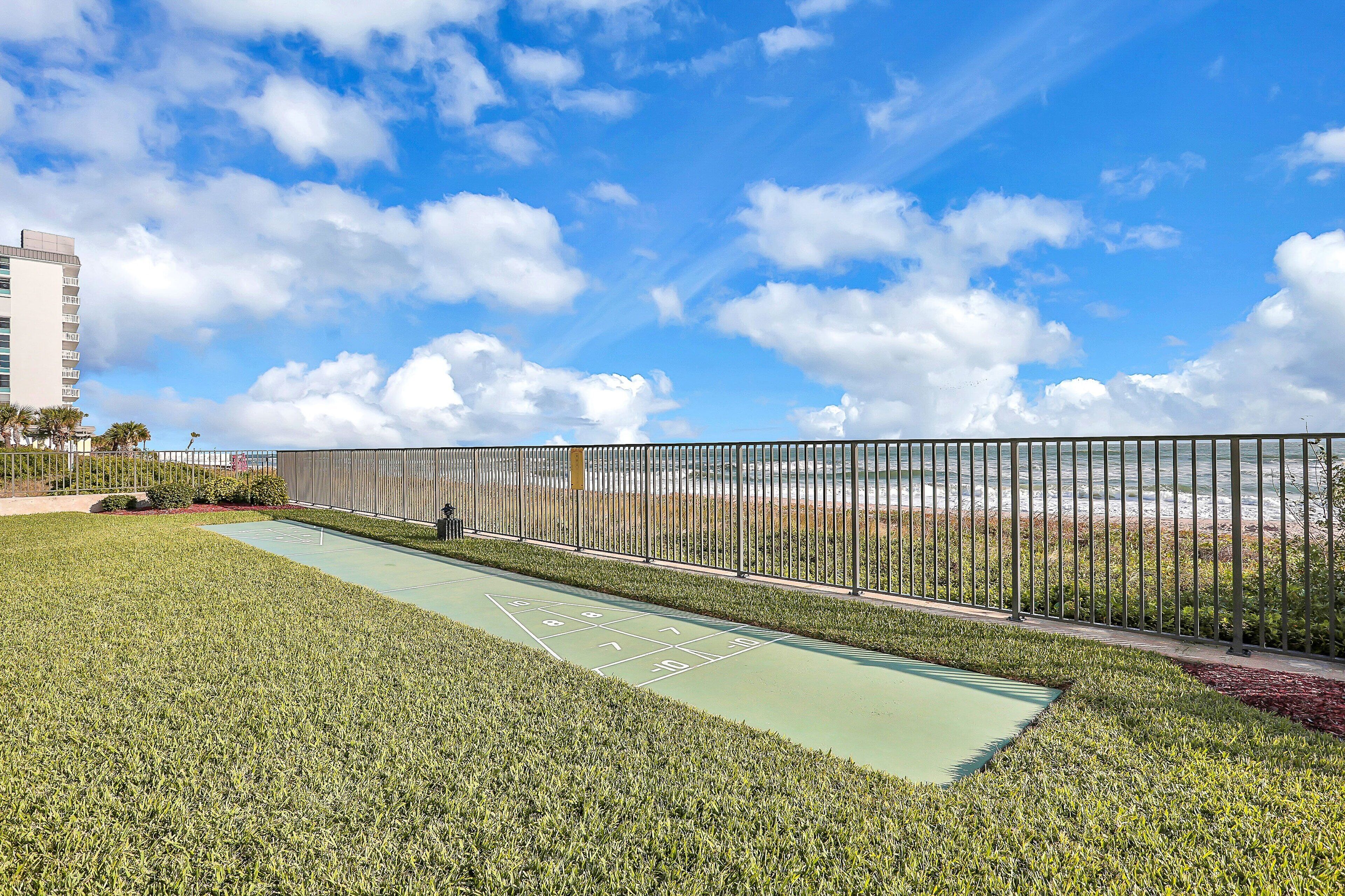 Un campo da shuffleboard verde all'aperto con recinzione in metallo, circondato da prato curato e vista libera sull'oceano.