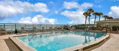 Ein rechteckiger Pool mit gefliestem Rand, umgeben von Liegestühlen und einem Zaun, mit Palmen und blauem Himmel im Hintergrund.
