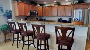 A kitchen with wooden cabinets, a stainless steel refrigerator, a microwave, and a granite countertop. There are four wooden bar stools and a potted plant. - NSB Malibu Condos 804-malibu 804 (New Smyrna Beach)