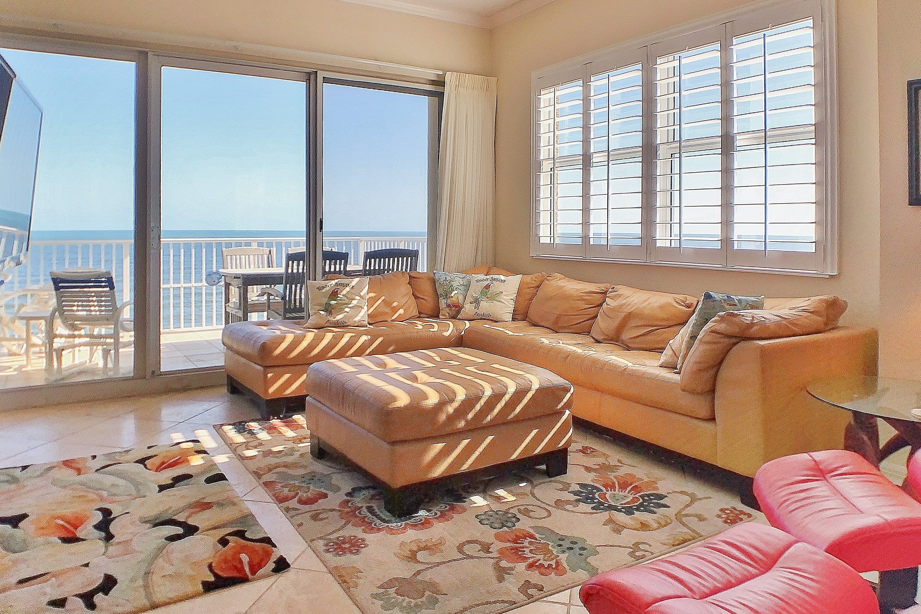 Un salon comprenant un canapé, un pouf et une table basse en verre. Une grande fenêtre, dotée de stores, offre une vue sur la mer.