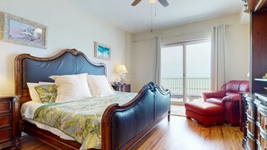 A spacious bedroom with a large bed, a ceiling fan, a red leather armchair, and a view of the outdoors. - NSB Malibu Condos 804-malibu 804 (New Smyrna Beach)