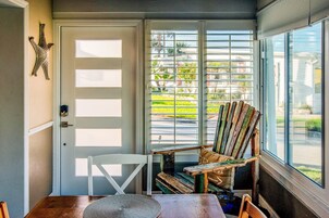 Una habitación con una mesa de comedor de madera, una silla blanca y una silla Adirondack de colores junto a un ventanal con persianas.