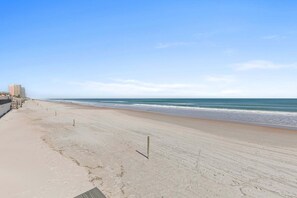 A beach with a clear sky, a sandy shore, and a building with multiple floors on the left. - Daytona Atlantico 32 (Daytona Beach)