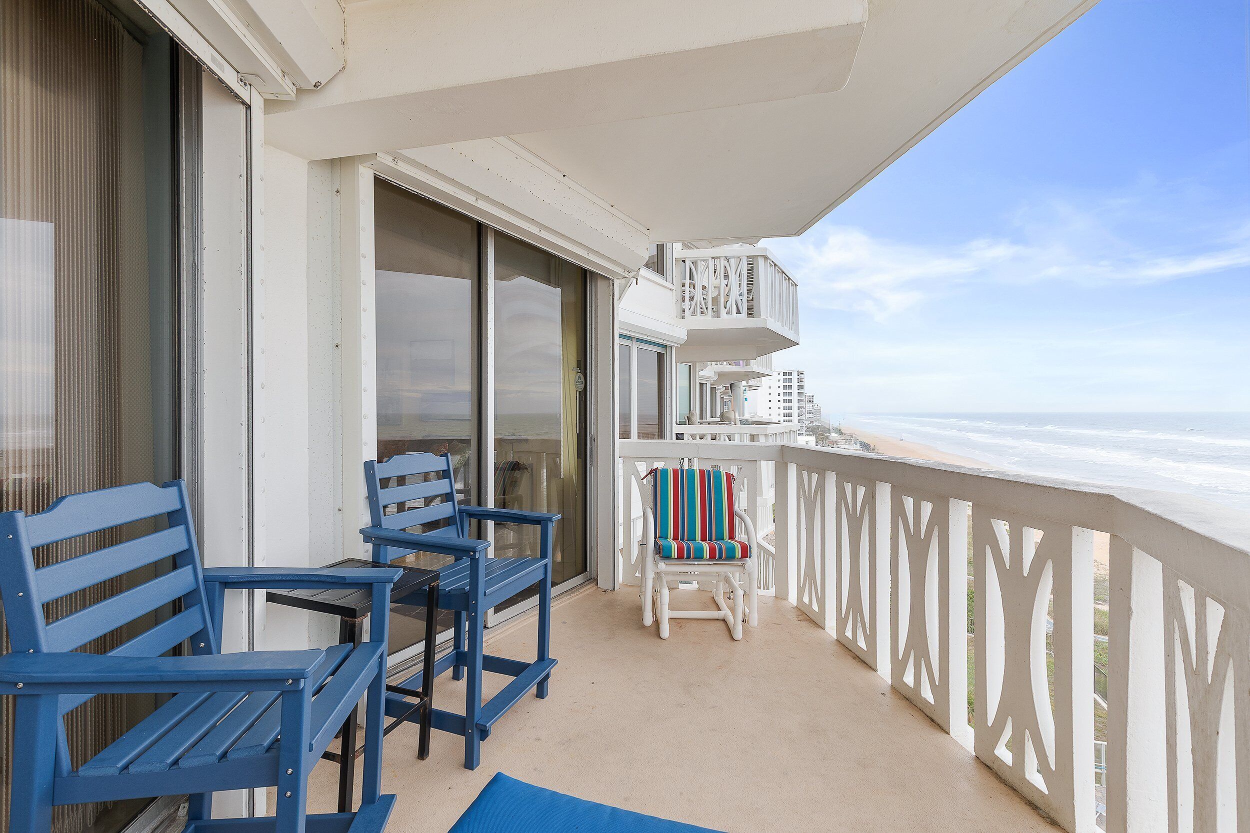 A balcony with two blue chairs and a white chair with a striped cushion, overlooking a beach.