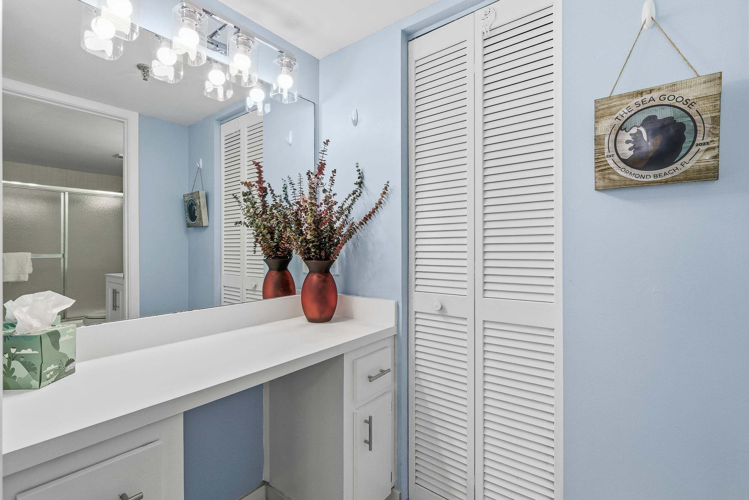 Een badkamer met een wit wastafelmeubel, twee koperen vazen met planten, een hanglamp en een bord met tekst aan de muur.