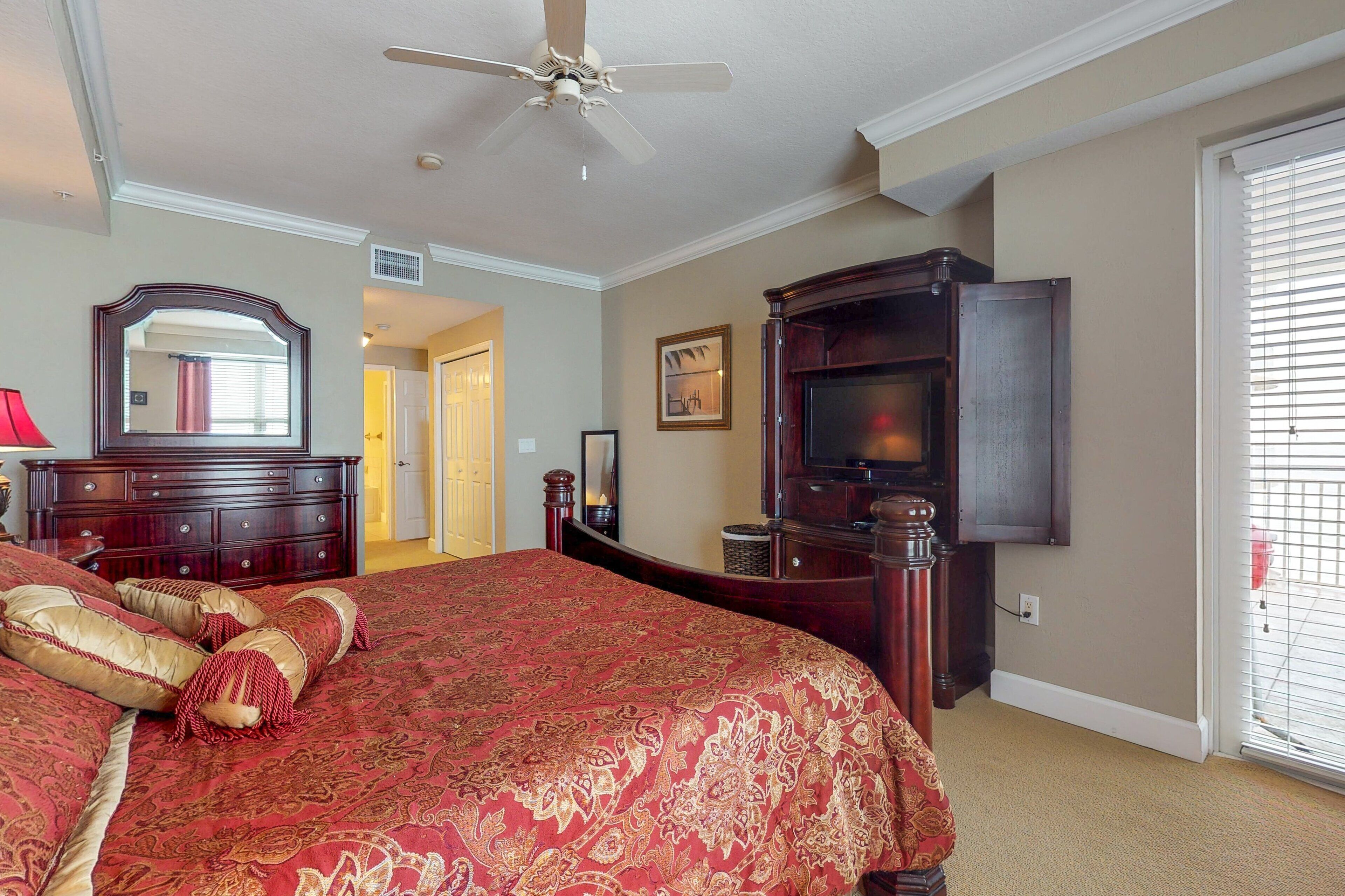 A bedroom with a large bed, a ceiling fan, a dresser, a television, and a door leading to another room.