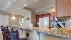 Una cocina con encimeras de granito, gabinetes de madera y un refrigerador. Se observan taburetes de bar y una cafetera.