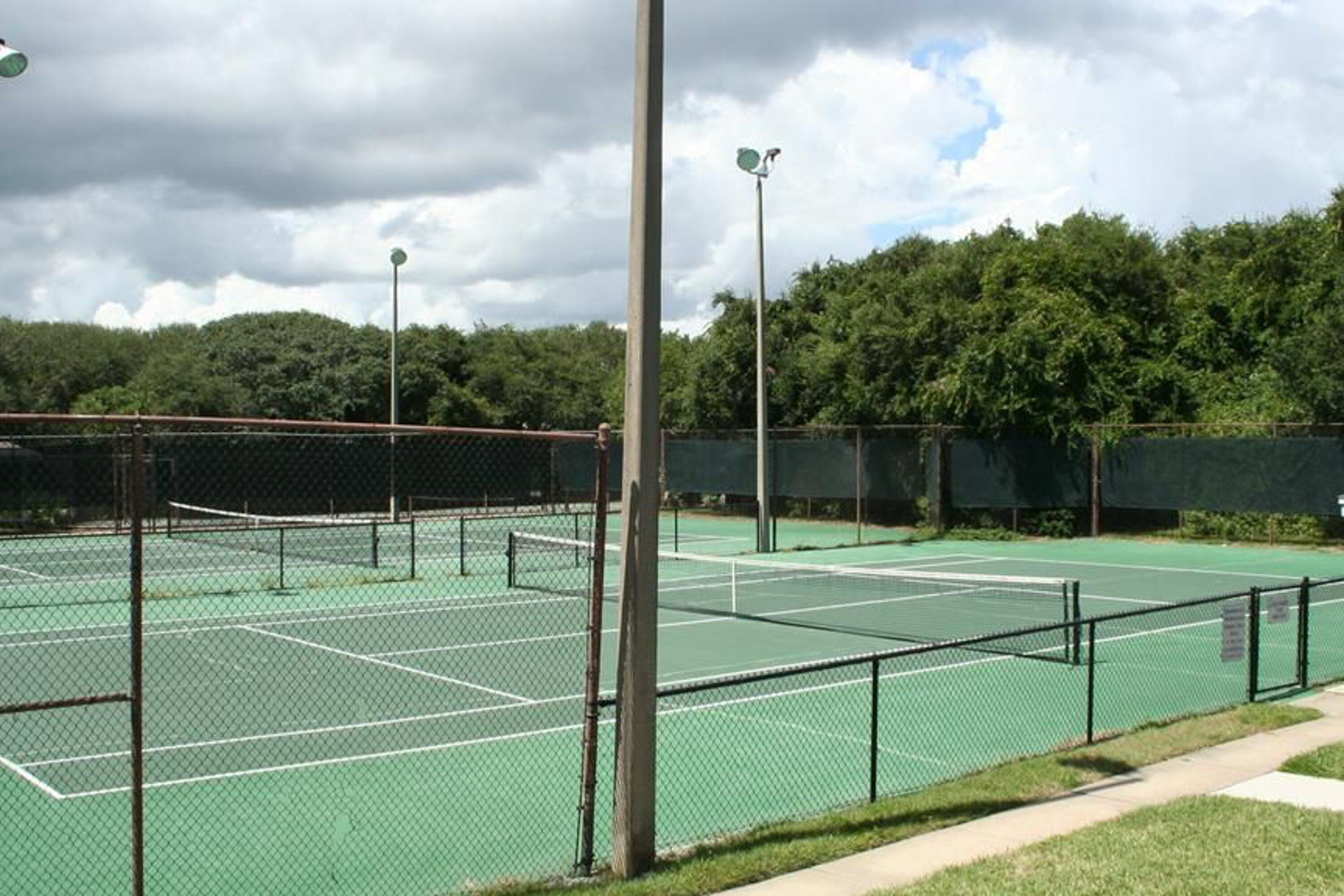 A tennis court with a net, surrounded by a chain-link fence, and trees in the background.