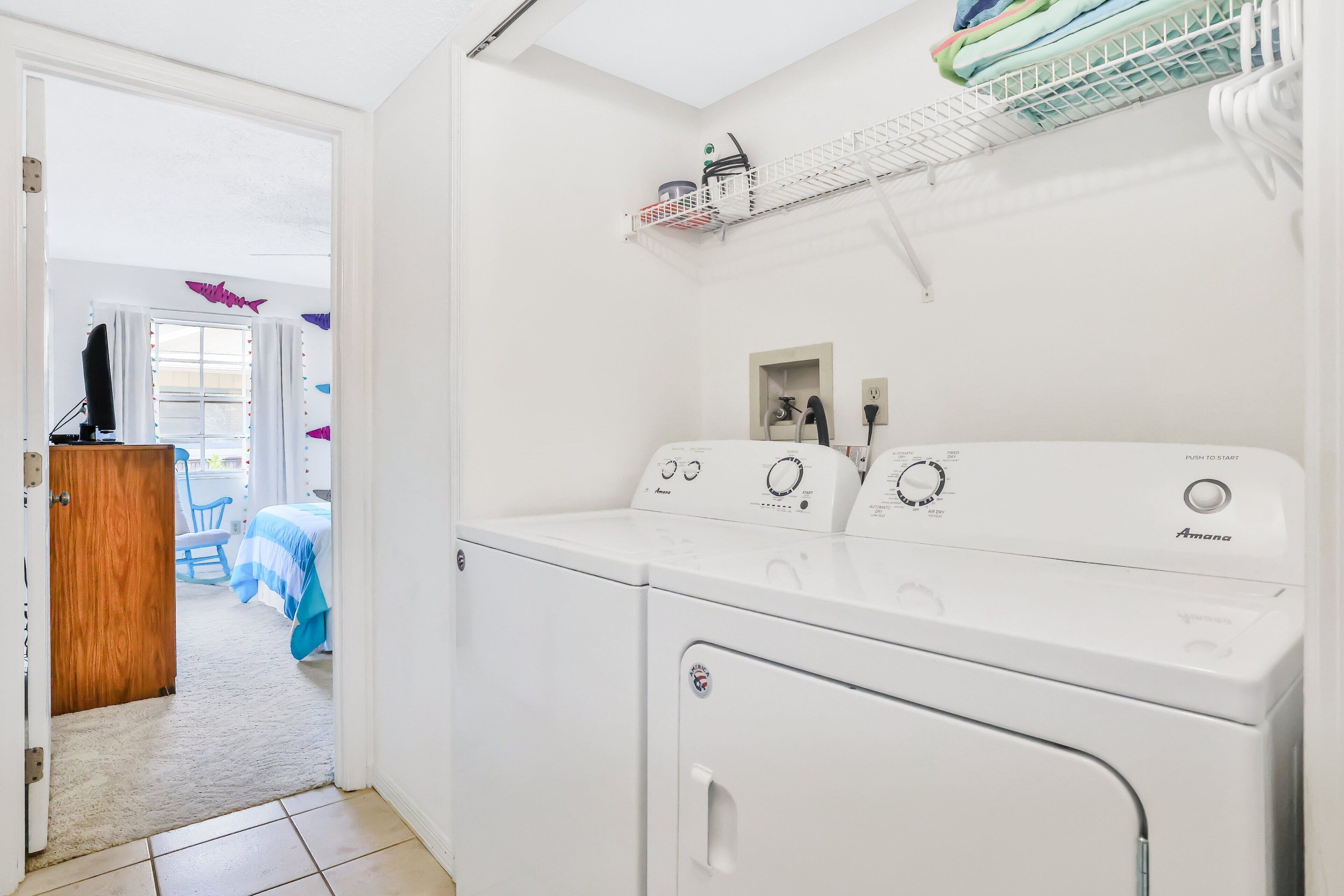 Een wasserette met een voorlader wasmachine en droger, een houten deur naar een slaapkamer, en een plank boven de droger met opgevouwen kleding en een wasmand.