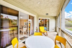 Uma área de varanda coberta com uma mesa redonda e cadeiras amarelas, portas de vidro deslizantes que dão para um espaço interno, e vista para o exterior.