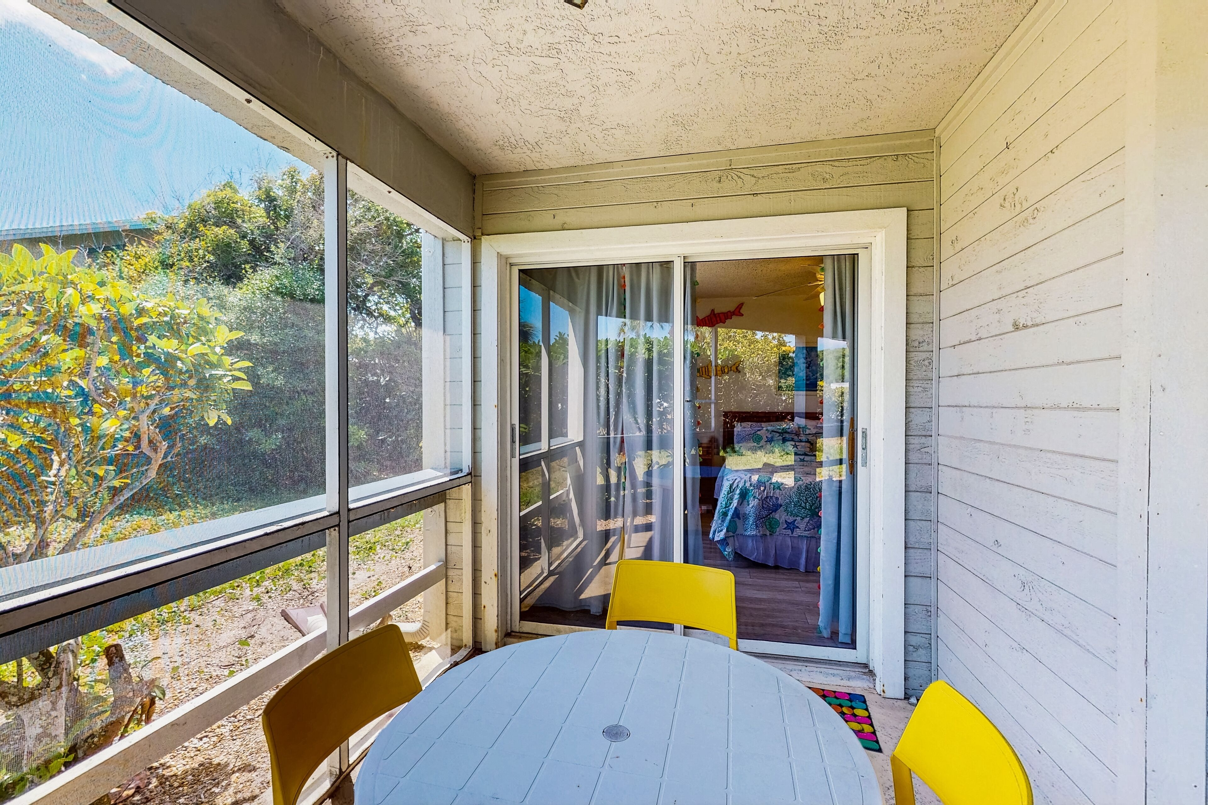 A covered patio with a table and chairs, a sliding glass door leading to an outdoor area with a table and chairs, and a view of trees and a house in the background.