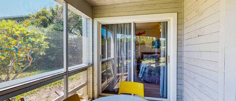 A covered patio with a table and chairs, a sliding glass door leading to an outdoor area with a table and chairs, and a view of trees and a house in the background.