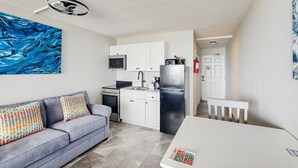 A modern kitchen with white cabinets, a stainless steel refrigerator, and a microwave. There is a gray sofa with patterned cushions and a white table with a placemat. - Daytona Pirates Cove 3270-salt Air Cove (Daytona Beach Shores)