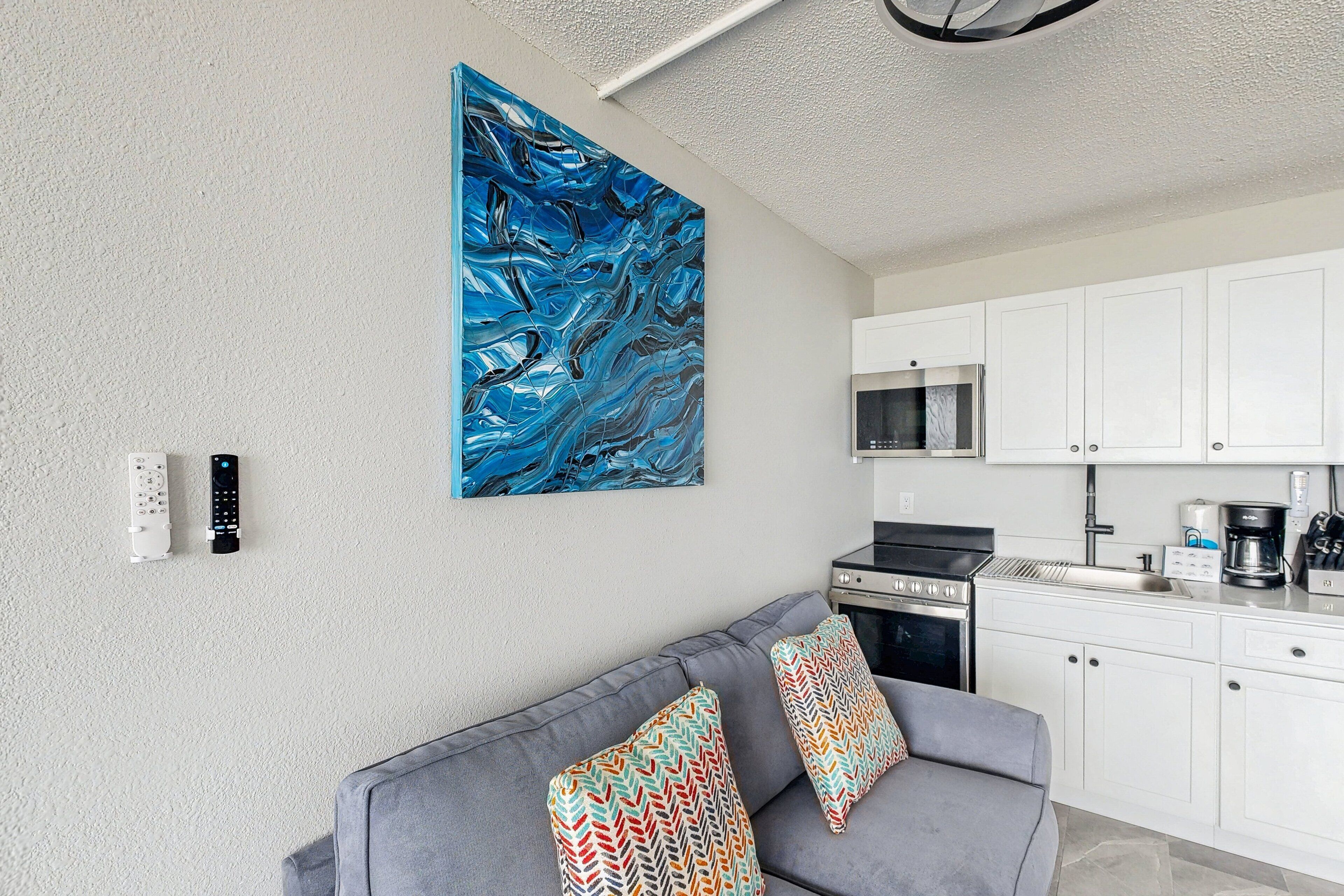 A modern living room with a grey sofa, colorful cushions, a large abstract painting, a kitchen with white cabinets, and a built-in oven.
