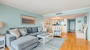 Ein Wohnzimmer mit einem grauen Ecksofa, einem weißen Couchtisch und einem blauen Teppich. Im Hintergrund ist eine Küche mit weißen Schränken und einer Pendelleuchte zu sehen.