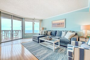 A living room with a sofa, coffee table, and a view of the ocean through large sliding doors. - Van Lee 1003 - Oceanview Beach Condo (Ormond Beach)