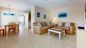 A spacious living room with a beige sofa, a wooden coffee table, and a dining area with a wooden table and chairs. - Nsb- Seaside Oasis (New Smyrna Beach)
