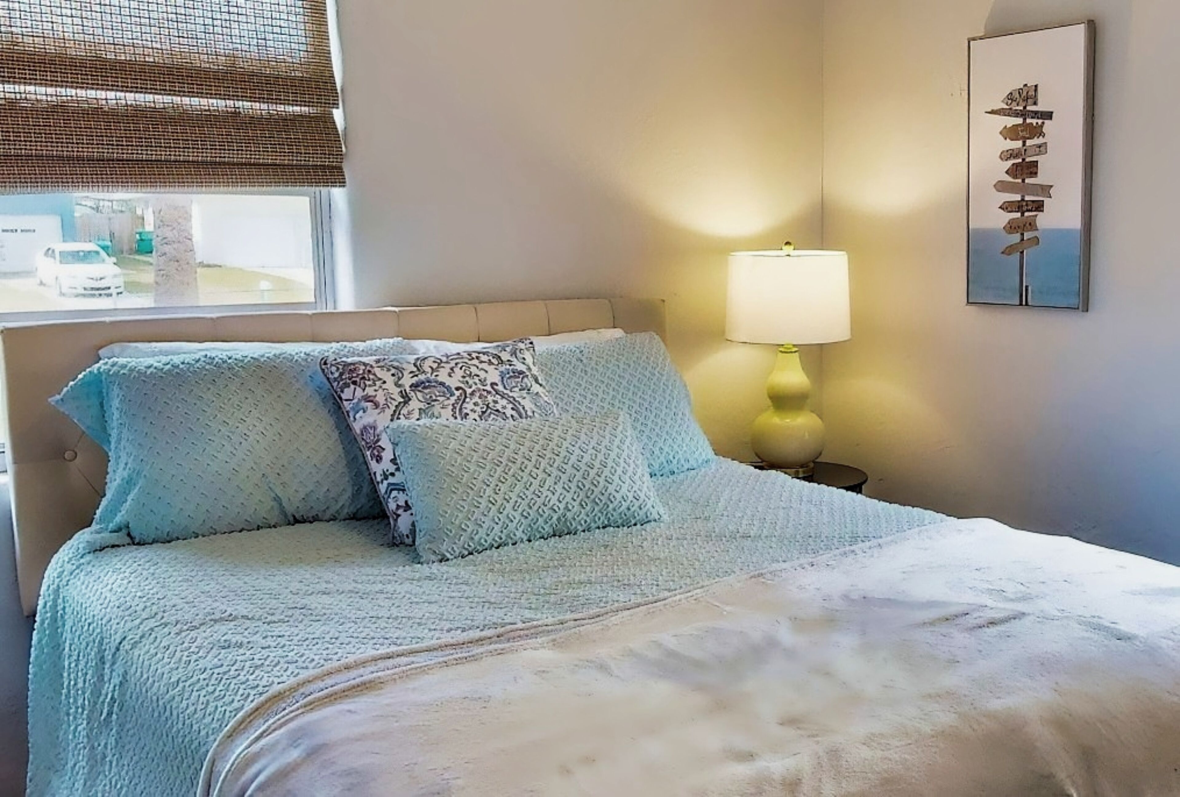 Un lit bien fait, avec une couverture bleu clair et des oreillers, une lampe de chevet et un tableau décoratif au mur.
