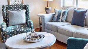 En stue med en mønstret lænestol, en sofa med puder, et lille bord med en lampe og en dekorativ glas skål på bordet.