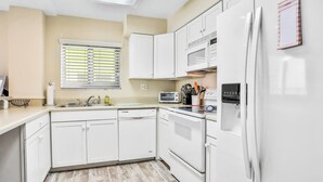 A modern kitchen with white cabinetry, a built-in microwave, a refrigerator, and a window with blinds. - Daytona Surfside Club 109- Blue Umbrella (Daytona Beach Shores)