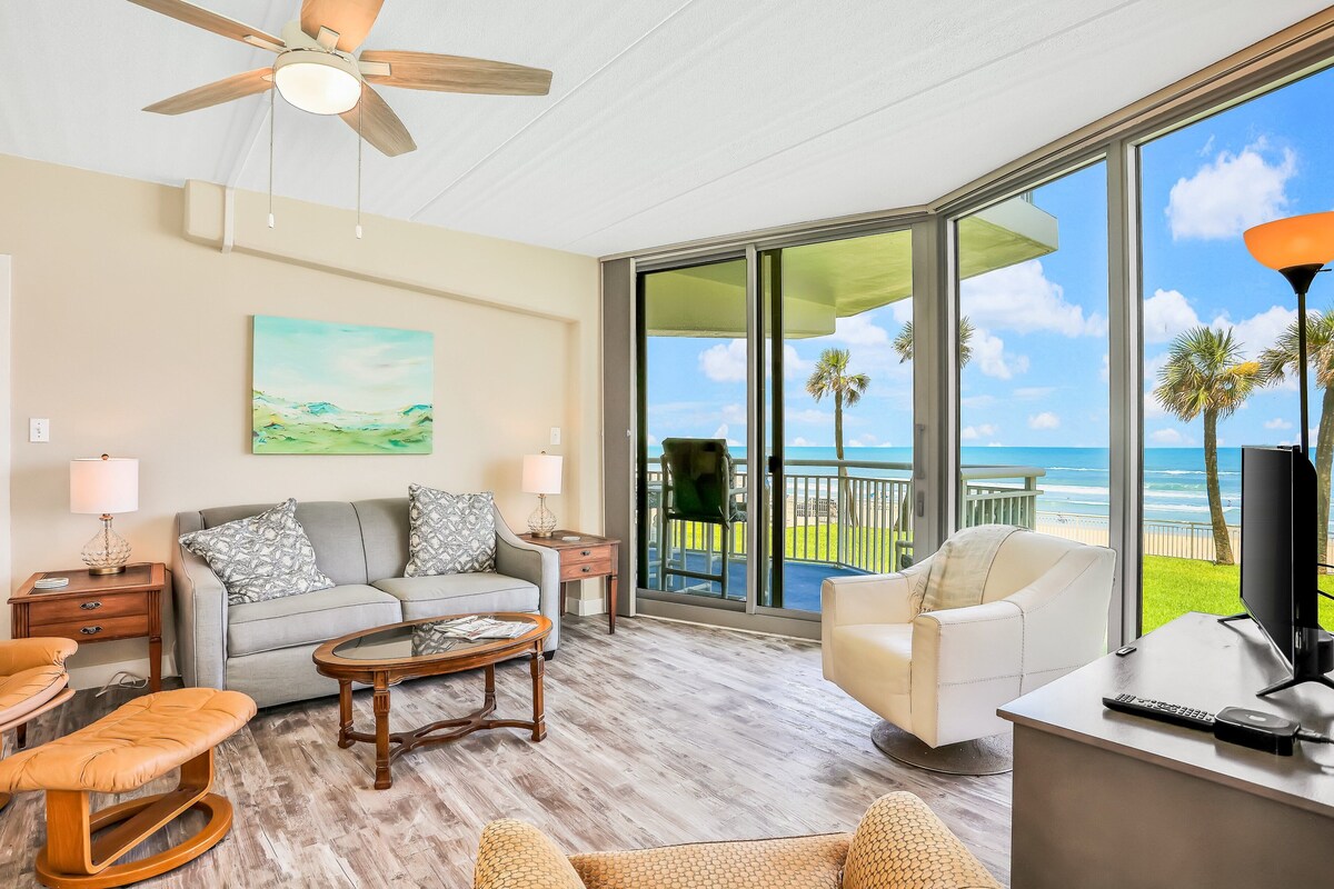 A spacious living room with a large window overlooking the ocean, a ceiling fan, and a comfortable seating area.