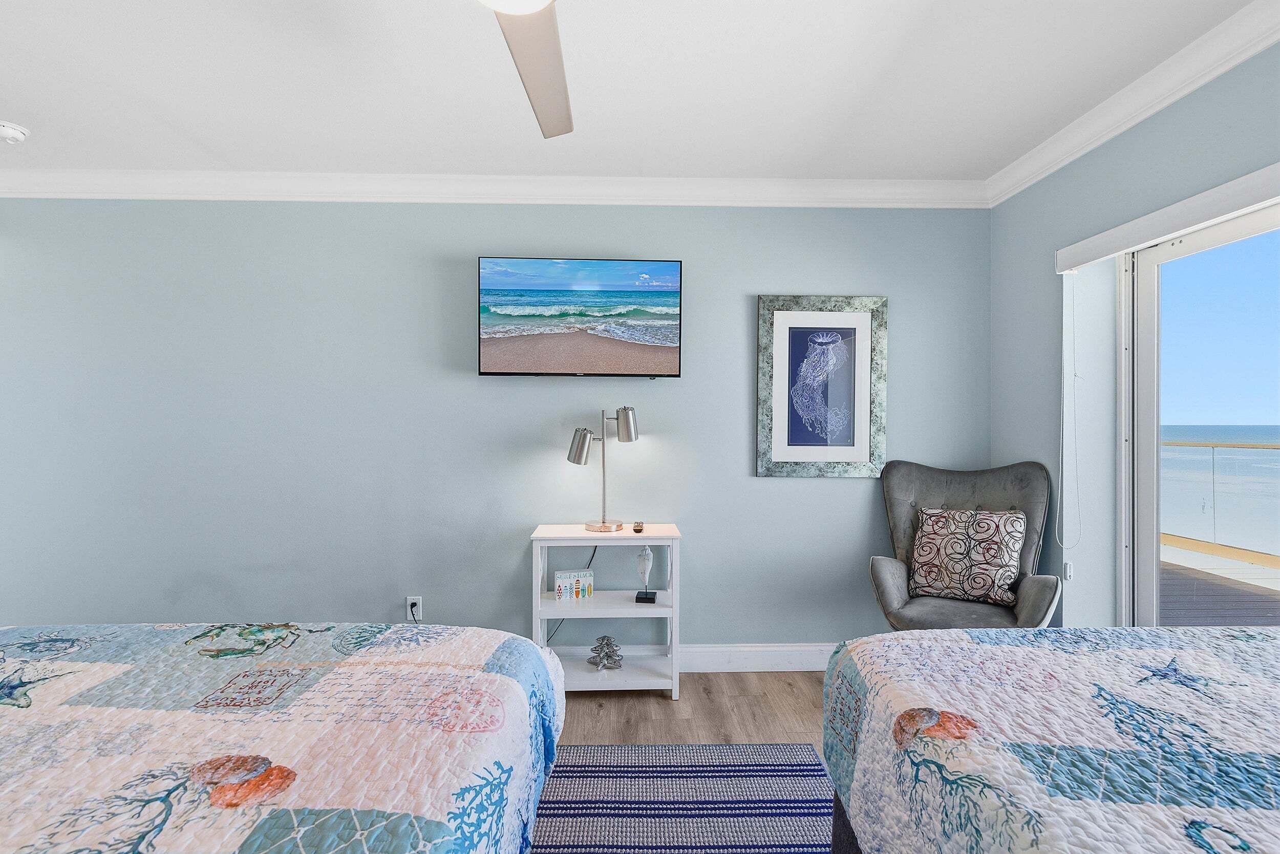 A bedroom with two beds, a TV, a nightstand, a chair, and a view of the beach.