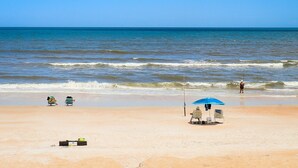 Ein Strand mit blauem Himmel, ruhiger See und wenigen Personen. Liegestühle und ein Sonnenschirm sind vorhanden.