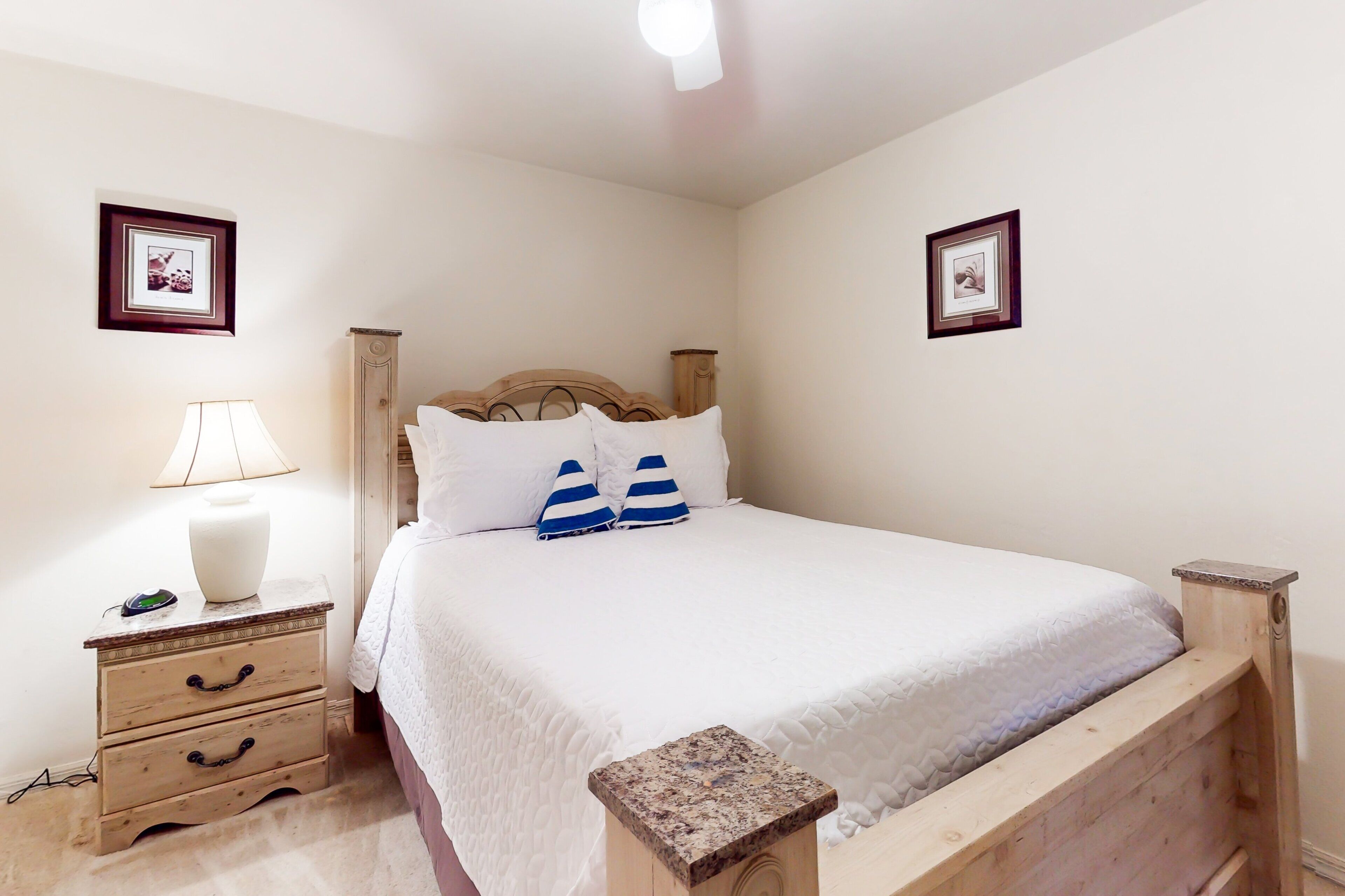 A neatly made bed with white bedding and blue striped pillows, a wooden headboard, a bedside table with a lamp, and two framed pictures on the walls.